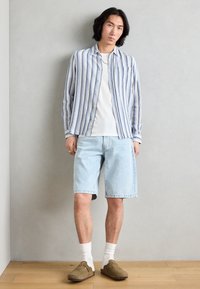Lyseblå denimshorts kombinert med en stripete langermet skjorte i blått og hvitt, akkompagnert av hvite sokker og brune slip-on sko.