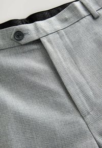 Pantaloni grigio chiaro su misura realizzati in tessuto strutturato con chiusura a bottone e pieghe sul davanti. La cintura è foderata in nero.