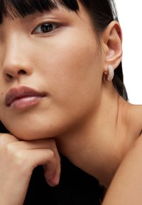 Rose gold hoop earrings featuring small white beads, with a smooth texture and a rounded design, worn by a person resting their chin on their hand.