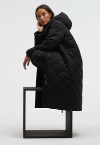 Schwarzer, gesteppter langer Mantel mit Kapuze, ausgestattet mit Reißverschluss und Tasche, präsentiert auf einem quadratischen grauen Sockel. Glatte Textur durchgehend.