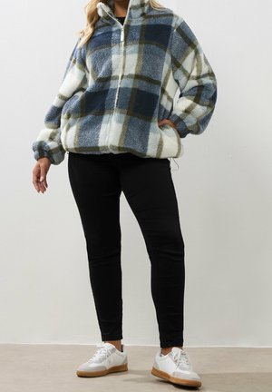 Kvinde iført en blå, hvid og olivengrønternet fleecejakke med sorte bukser og hvide sneakers, stående med den ene hånd i lommen.