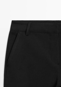 Gros plan sur la ceinture d’un pantalon noir avec passants de ceinture et poche fendue sur le côté gauche, sur fond blanc.