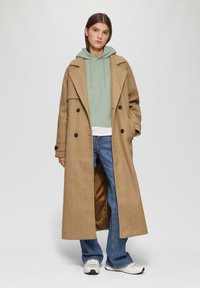 Brauner Wollmischungs-Mantel im Trenchcoat-Stil mit Doppelreihiger Knopfleiste, kombiniert mit einem hellgrünen gerippten Hoodie und blauen Schlagjeans. Weiße Sneakers sind sichtbar.