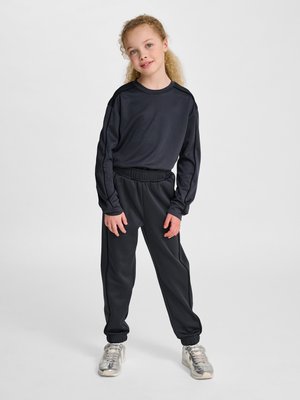 Jong meisje met krullend haar staat met een zwarte top met lange mouwen, zwarte joggingsbroek en zilveren sneakers tegen een effen achtergrond.