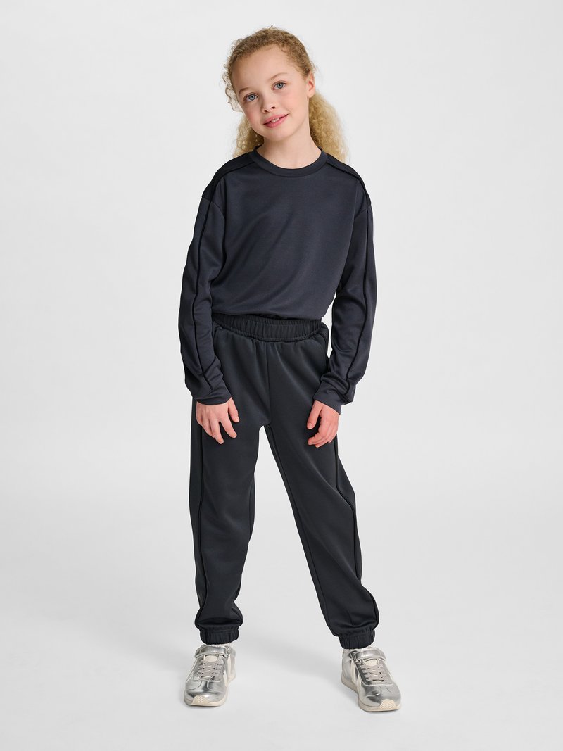 Ung pige med krøllet hår står iført en sort langærmet top, sorte joggingbukser og sølvfarvede sneakers mod en ensfarvet baggrund.