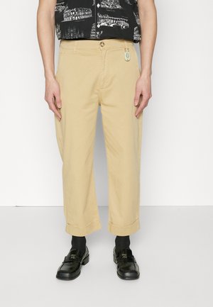 Homme portant un pantalon beige retroussé, des chaussettes noires et des mocassins noirs, se tenant contre un fond blanc avec les mains détendues sur les côtés.