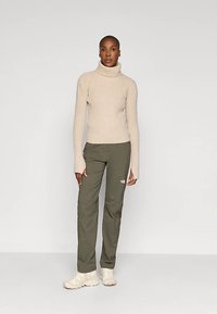 Femme portant un pull col roulé beige, un pantalon vert olive avec un logo sur la cuisse et des baskets claires, debout contre un fond uni.