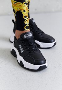 Zapatillas deportivas negras de material sintético con suela blanca. Sus características incluyen cordones, marca en la lengüeta y un diseño voluminoso.
