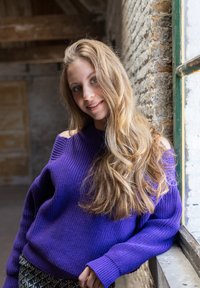 Paarse gebreide trui met een brede halslijn en ribbelstructuur, gecombineerd met een patroontaille. Lange golvende haren omlijsten het gezicht. Natuurlijk licht vanuit een raam.