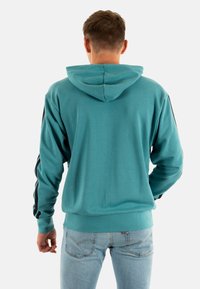 Felpa turchese con cappuccio posteriore, tessuto testurizzato, strisce laterali nere a contrasto e polsini a coste, abbinata a jeans azzurri chiari.