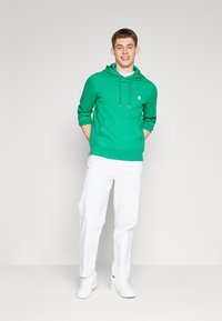 Grüne Kapuzenjacke mit Vordertasche, Kordelzugkapuze und Logo. Kombiniert mit weißen Hosen und weißen Turnschuhen, vor einem hellen Hintergrund stehend.
