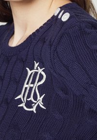 Mörkblå stickad tröja med kabelstickat mönster, med en vit broderad logotyp och tre vita knappar vid axelsömmen.