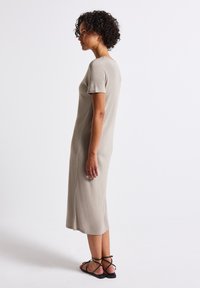 Šaty do kolen světle béžové barvy vyrobené z texturované látky, s krátkými rukávy a kulatým výstřihem, doplněné černými sandály na tenkých řemíncích.