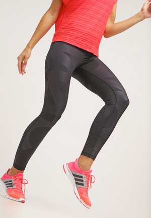 Leggings de sport noires avec un motif géométrique, associées à un haut texturé rouge et des chaussures de course roses vives. Jambes dans une pose dynamique.