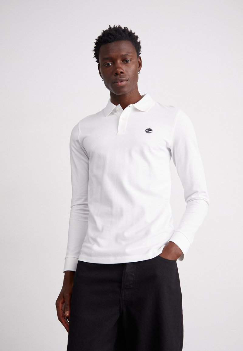 Vīrietis valkā baltu garo piedurkņu polo krekli ar mazu logotipu uz krūtīm un melnām biksēm, stāv ar vienu roku kabatā.