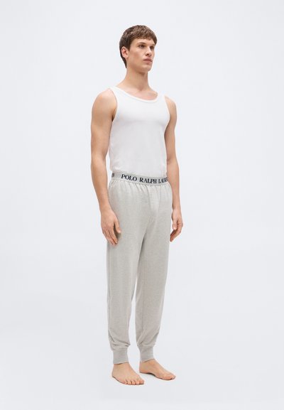 Νεαρός άνδρας όρθιος, ξυπόλυτος, φοράει λευκή φανέλα και ανοιχτό γκρι φόρμα με ζώνη μέσης "Polo Ralph Lauren", απλό ανοιχτόχρωμο φόντο.