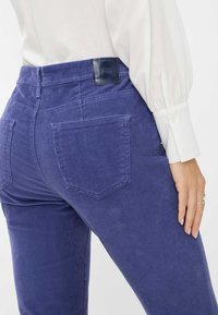 Pantaloni in velluto a coste viola con superficie testurizzata, dotati di tasche posteriori e un'etichetta in tessuto blu navy sulla vita. Il modello indossa una camicia bianca.