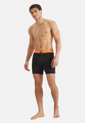 Oberkörperfreier Mann steht barfuß und trägt schwarze Boxershorts mit einem orangefarbenen Bund, auf dem ein schwarzes Adidas-Logo zu sehen ist.