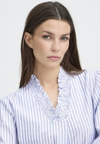 Camicia bianca con strisce blu, caratterizzata da un collo arricciato, maniche a sbuffo e una vestibilità comoda. Tessuto morbido con una texture leggera.