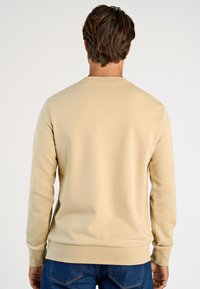 Beige huppari pitkillä hihoilla, pyöreällä kaula-aukolla ja ribbausmateriaalilla varustetuilla hihoilla. Ominaisuuksiin kuuluu sileä rakenne ja rento leikkaus, joka on yhdistetty tummansinisiin farkkuihin.