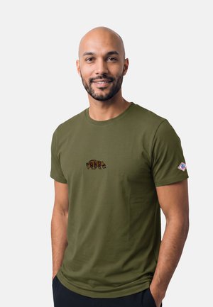 Uomo calvo sorridente con la barba che indossa una maglietta verde oliva con un piccolo logo di orso sul petto e una toppa sulla manica sinistra.