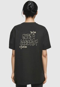 T-shirt oversize noir en coton, avec un design graphique texturé en texte beige clair : "Cet assistant de Cupidon cher, c'est votre tour" au dos.