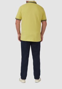 Kurzarm-Poloshirt in hellgelb mit dunkelgestreiftem Kragen, kombiniert mit marineblauen Hosen und hellen Schuhen.