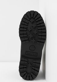 Schwarze Gummisohle mit tiefem Profil und Kreuzmustern, mit einem Logo und dem Text "Timberland" in der Nähe der Ferse, auf einem weißen Hintergrund angewinkelt.