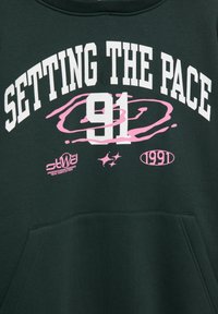 Felpa verde con testo bianco "SETTING THE PACE" sopra il numero 91, accentuata da grafiche a spirale rosa e loghi aggiuntivi in basso.