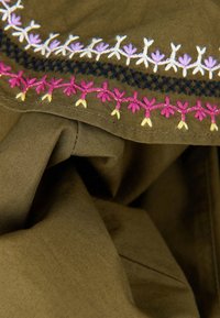 Tessuto marrone con ricami intricati che presentano motivi floreali rosa, viola, bianchi e neri lungo il bordo. Materiale morbido e texturizzato.