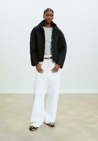Veste noire matelassée avec col haut, fermetures à pression, portée sur un haut à manches longues gris, associée à un jean large blanc et des chaussures à motifs.