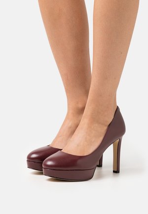 Paire de jambes portant des chaussures à plateforme marron foncé à talons hauts sur fond blanc, les pieds rapprochés et légèrement inclinés.