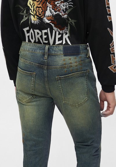 Personne portant un jean bleu délavé avec de petits motifs en croix près de la ceinture et un t-shirt noir avec un tigre rugissant et le texte "FOREVER".