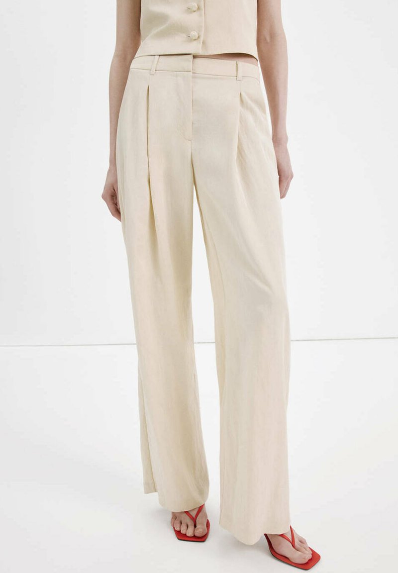 Pantalon beige taille haute à jambes larges associé à un top sans manches assorti et des sandales rouges à bout carré sur une personne debout.