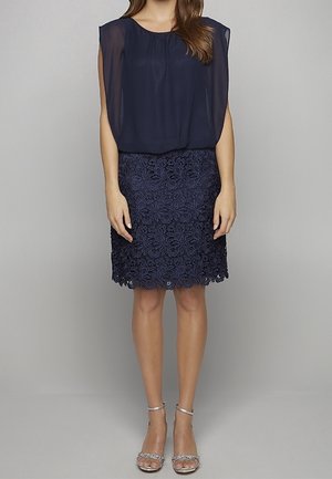 Navy blue two-piece outfit featuring a sheer draped top and a fitted lace skirt with floral patterns and scalloped hem. Silver ankle-strap heels.