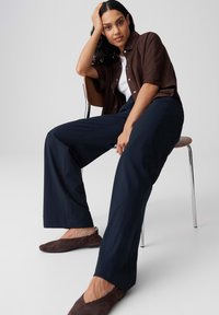 Femme aux cheveux bouclés foncés assise sur une chaise, portant une chemise marron, un haut blanc, un pantalon large bleu marine et des chaussures plates marron, sur un fond uni.