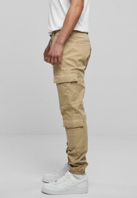Beige Cargo-Hosen mit mehreren Taschen, schmal zulaufenden Bündchen und einem Kordelzug. Getragen mit weißen Sneakern. Seitliche Ansicht.