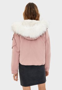 Kort rosa jacka med en vit fuskpälsluva, elastiska muddar och en figursydd design. Tyget är slätt och något glansigt.