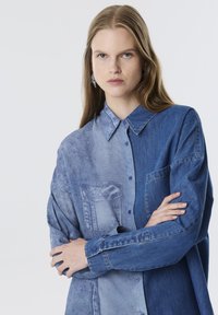 Camisa de mezclilla de botones con un diseño de patchwork en tonos de azul claro y oscuro, con diferentes texturas y un detalle de bolsillo en el pecho.