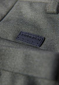Grüner Stoff mit einer strukturierten Oberfläche, ausgestattet mit einem dunklen Lederetikett, auf dem "LINDBERGH WHITE" prägedruckt ist. Nahaufnahme zeigt die Oberflächendetails.