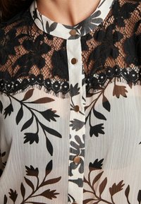 Schwarze Spitzenbluse mit floralen Motiven auf einem transparenten weißen Hintergrund. Verfügt über einen runden Kragen, eine Knopfleiste und filigrane Blattmuster in dunklen Tönen.
