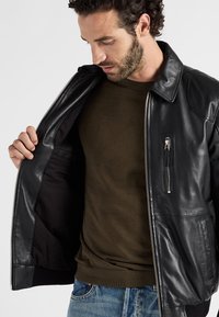 Schwarze Lederjacke mit Reißverschluss, ausgestattet mit einer Brusttasche und gerippten Bündchen, getragen über einem olivgrünen Rundhalsausschnitt-Shirt.