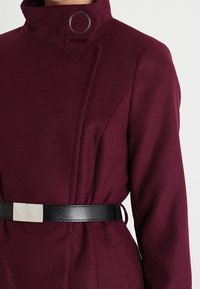 Burgundský kabát s vysokým límcem, který má stříbrný kroužkový detail a černý kožený pásek se čtvercovou sponou. Hladká, strukturovaná tkanina.