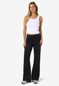 Mlada ženska z dolgimi blond lasmi, oblečena v bel brezrokavnik in črne široke kavbojke ter srebrne superge, stoji.