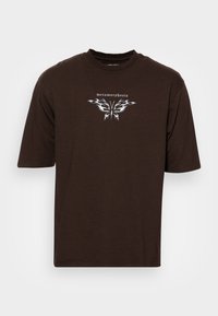 T-shirt en coton marron à manches courtes, ornée d'un design de papillon argenté métallique et du mot "métamorphose" au-dessus.