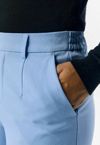 Hellblaue maßgeschneiderte Hose aus glattem Material, mit einem strukturierten Bund, seitlichen Taschen und elastischen Akzenten in der Taille.