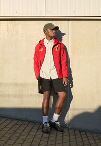 Červená bunda s kapucňou a logom, béžová košeľa s gombíkmi, čierne šortky s emblémom, zelená čiapka, biele ponožky a čierne tenisky. Pozadie z betónu.