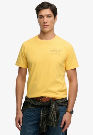 Man draagt een geel Superdry T-shirt, blauwe jeans, een bruine riem, een geruit overhemd om de taille gebonden en een polshorloge, staand tegen een witte achtergrond.