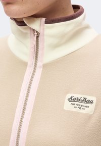 Gros plan d'une veste polaire beige avec une fermeture éclair à l'avant et un patch "Karitraa For Her By Her" sur la poitrine.