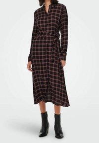 Eine Frau trägt ein schwarzes und rotes kariertes langärmliges Midikleid mit einem Taillengürtel und schwarzen Stiefeln bis zur Knöchel, während sie vor einem weißen Hintergrund steht.
